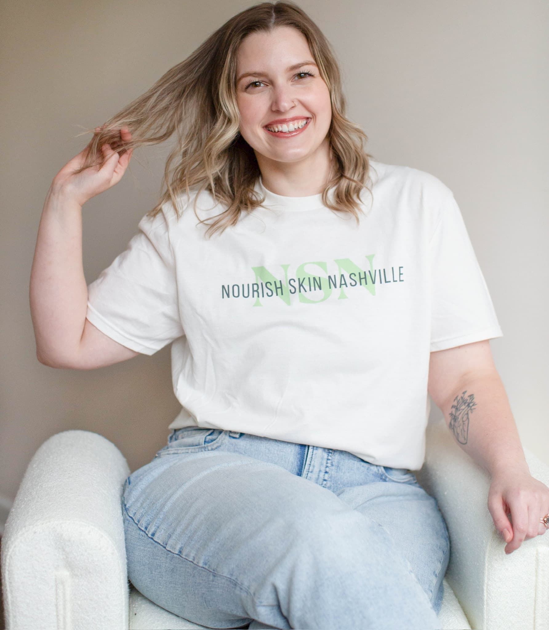 Katie Wyllia seated in a white armchair, lifting her hair with one hand and laughing
