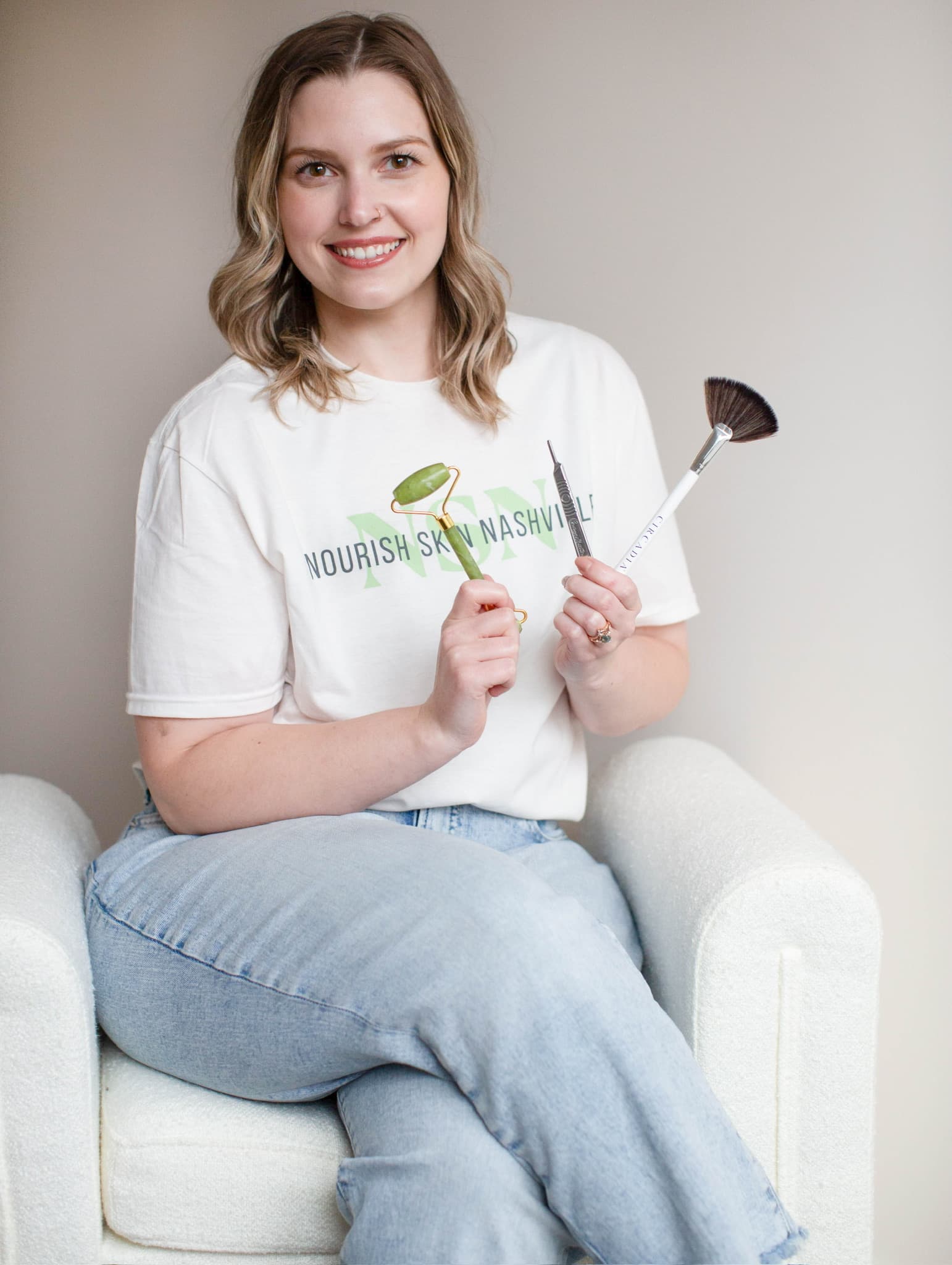 Katie Wyllia seated in a white armchair, holding a jade roller in one hand and a fan brush and spatula in the other, smiling