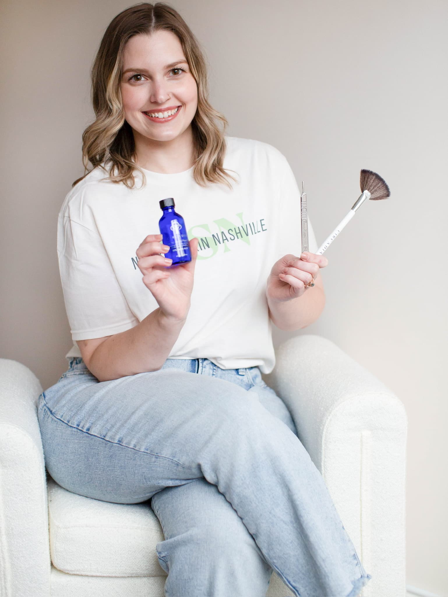 Katie Wyllia, licensed esthetician at Nourish Skin Nashville, seated in a white armchair, holding a blue serum bottle and a fan brush