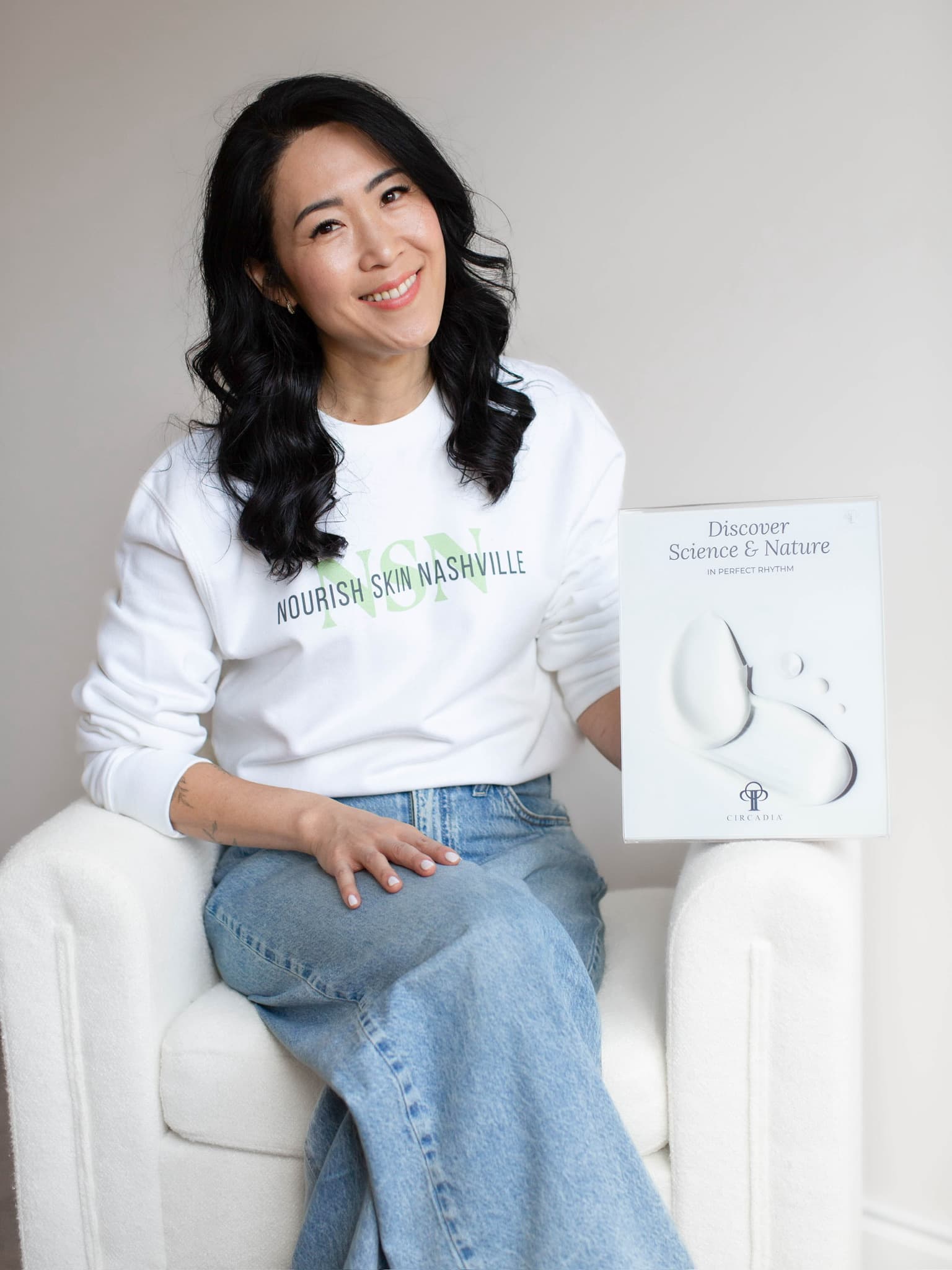 Kay Mason seated in a white armchair beside a Circadia skincare book propped on the chair arm, smiling