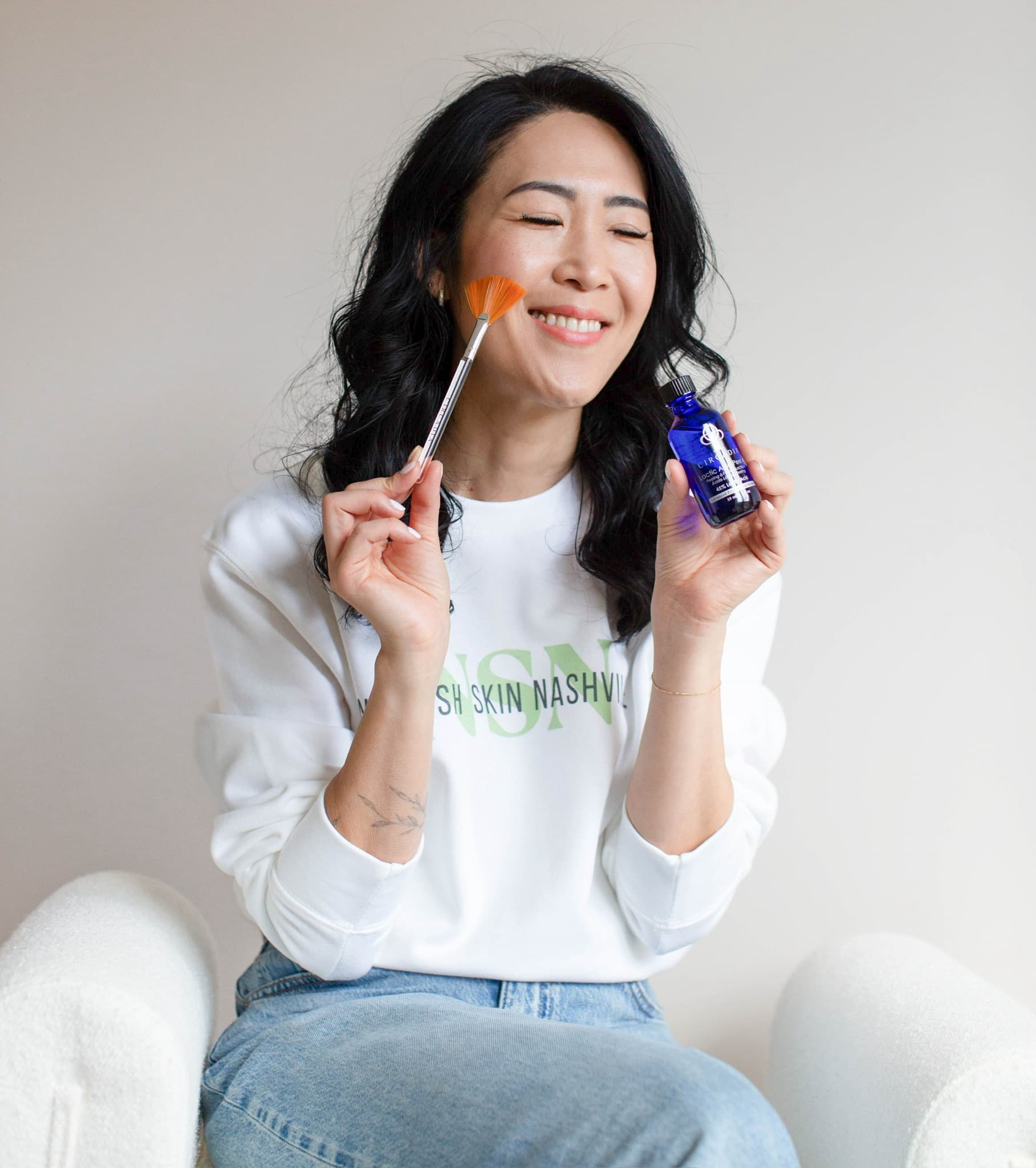 Kay Mason laughing with her eyes closed, a fan brush at her cheek and a serum bottle raised