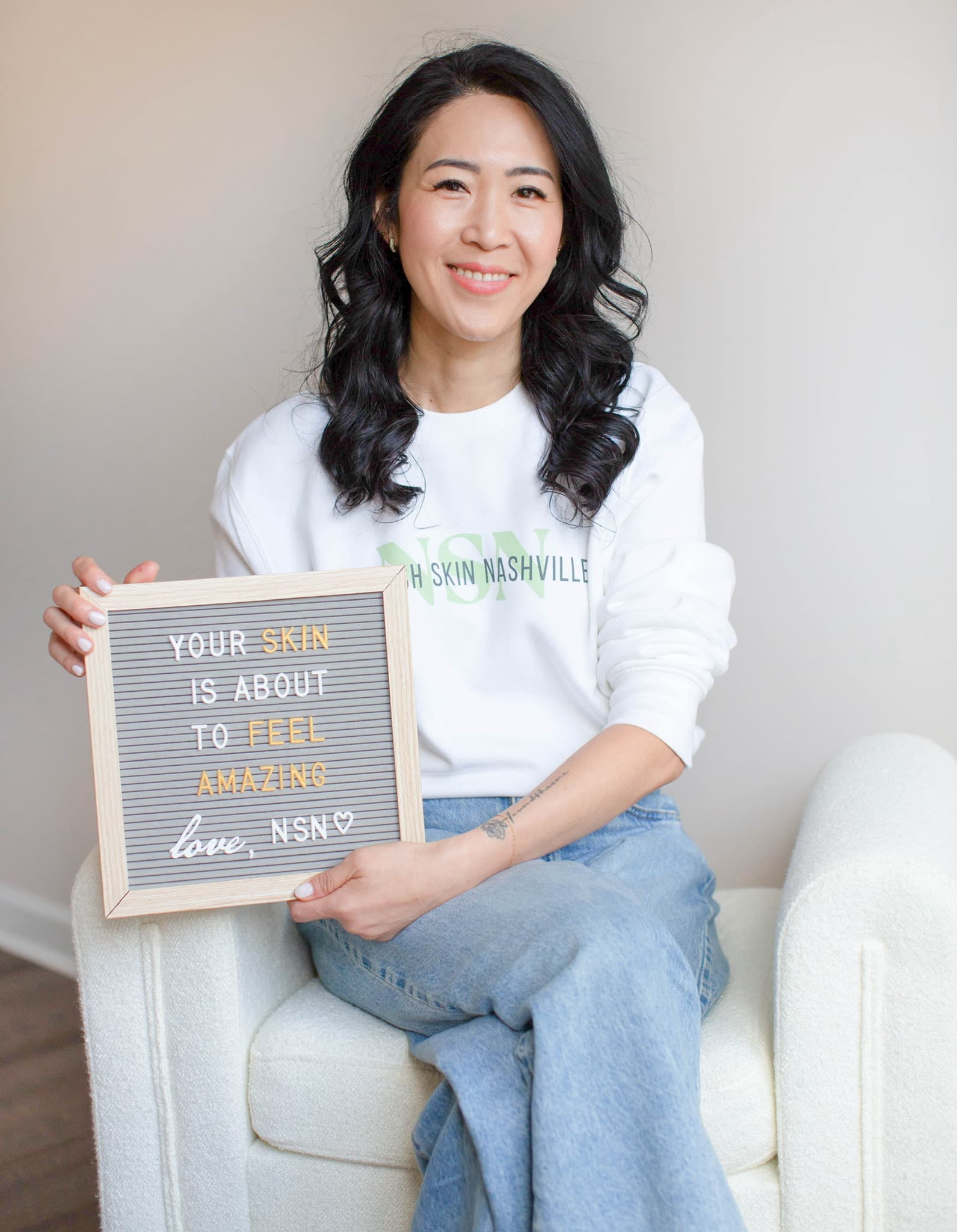 Kay Mason seated in a white armchair holding the studio's felt letterboard, smiling