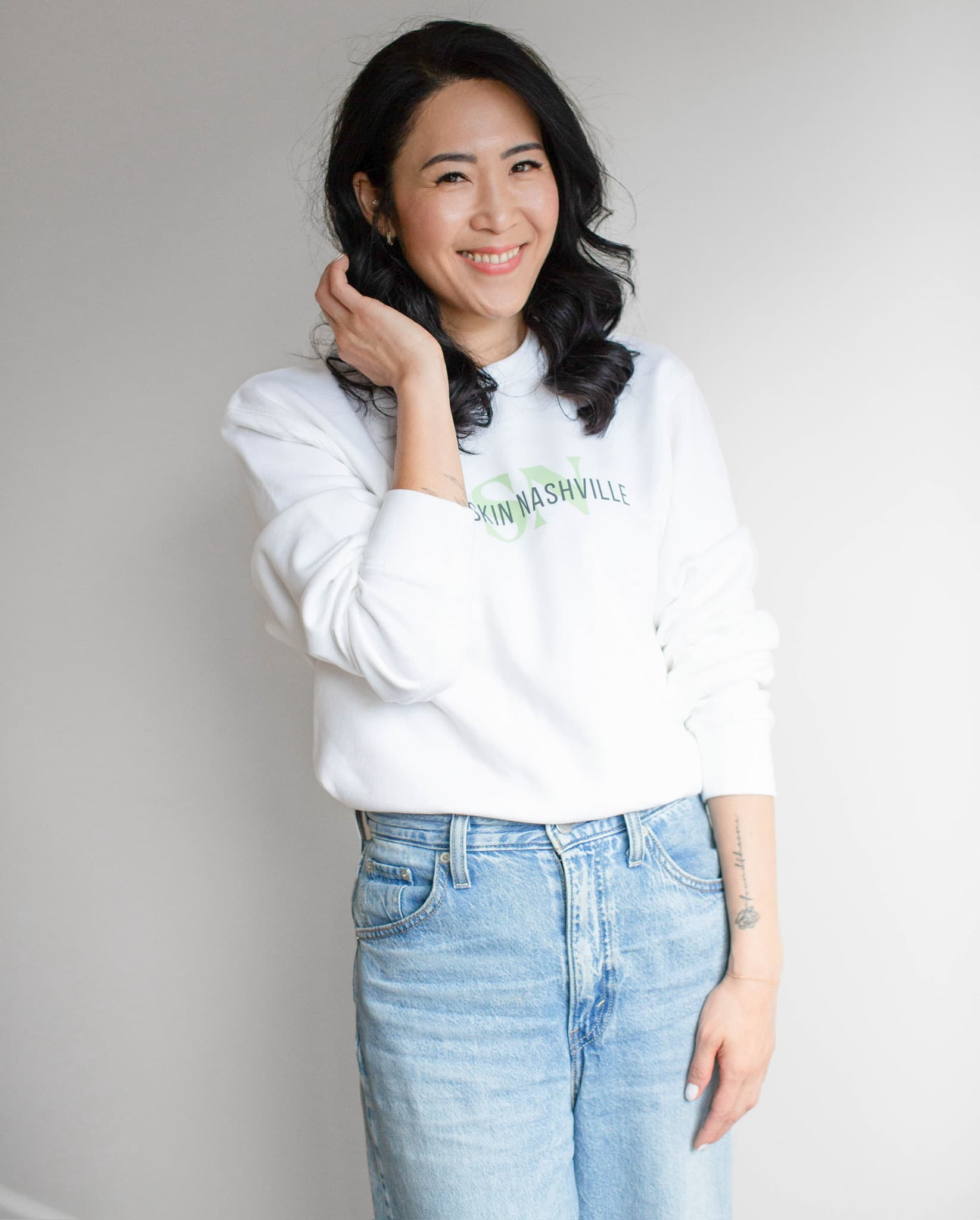Kay Mason standing against a grey wall, one hand at her hair, smiling