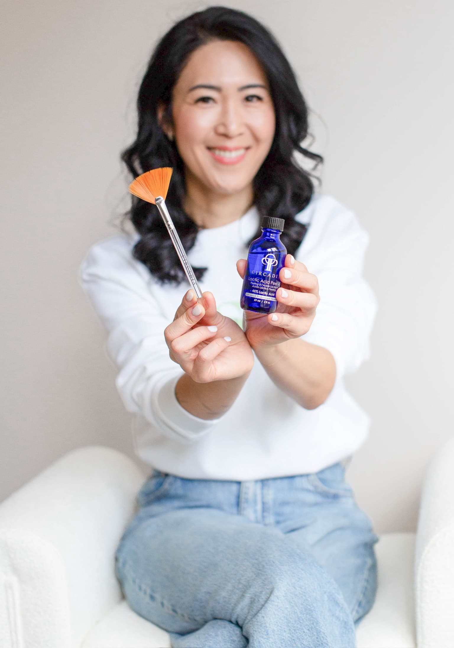 Kay Mason, licensed esthetician at Nourish Skin Nashville, seated in a white armchair, holding a fan brush and a blue serum bottle