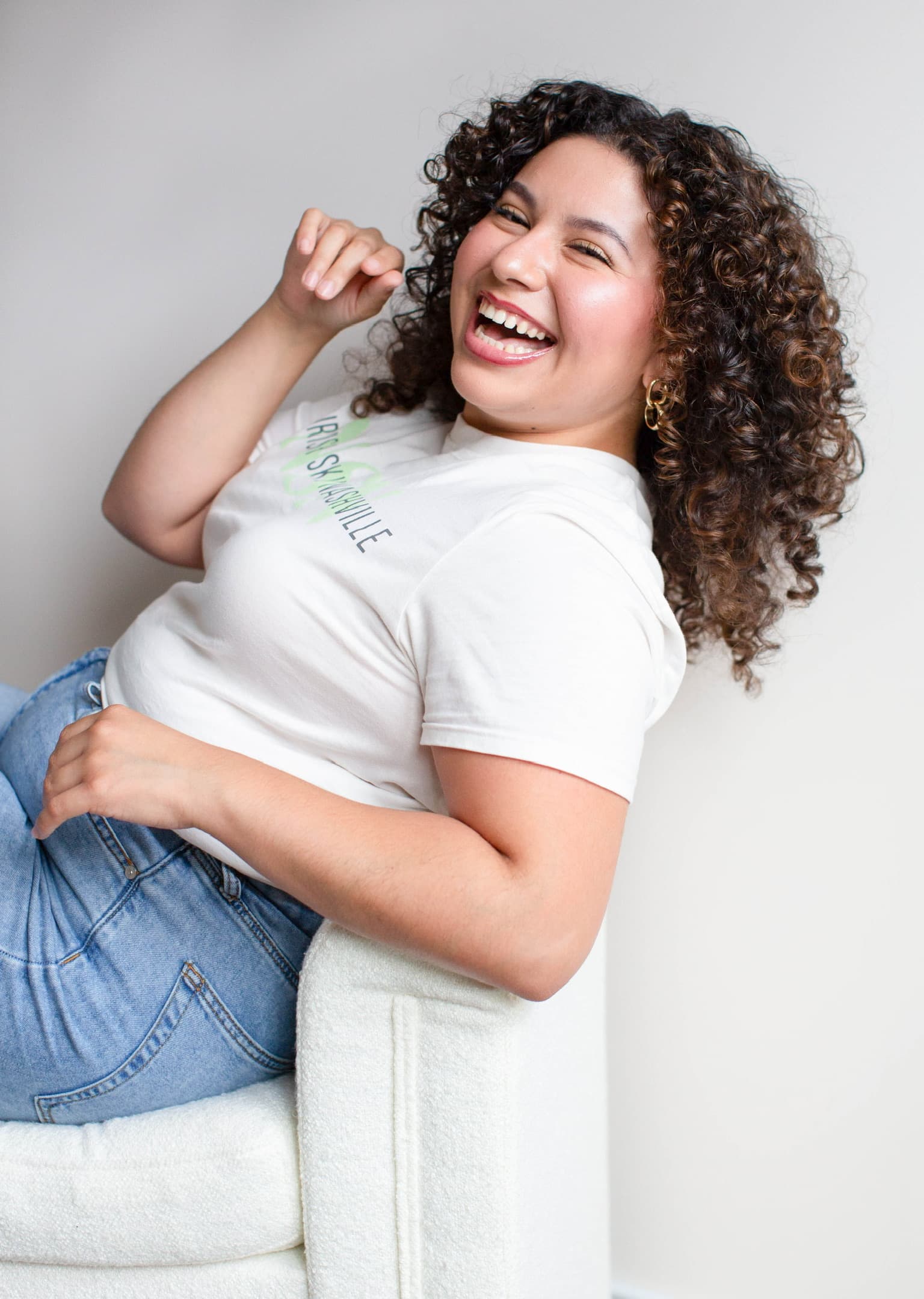 Roselyn Hernandez reclined sideways in a white armchair, one arm raised, laughing