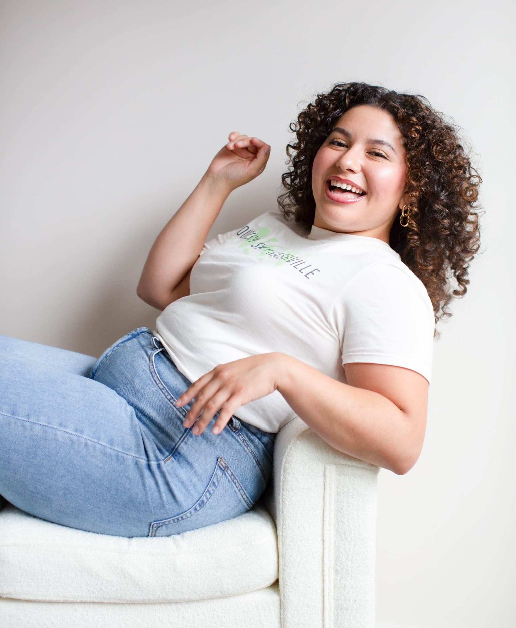 Roselyn Hernandez reclined sideways in a white armchair, arm raised, laughing