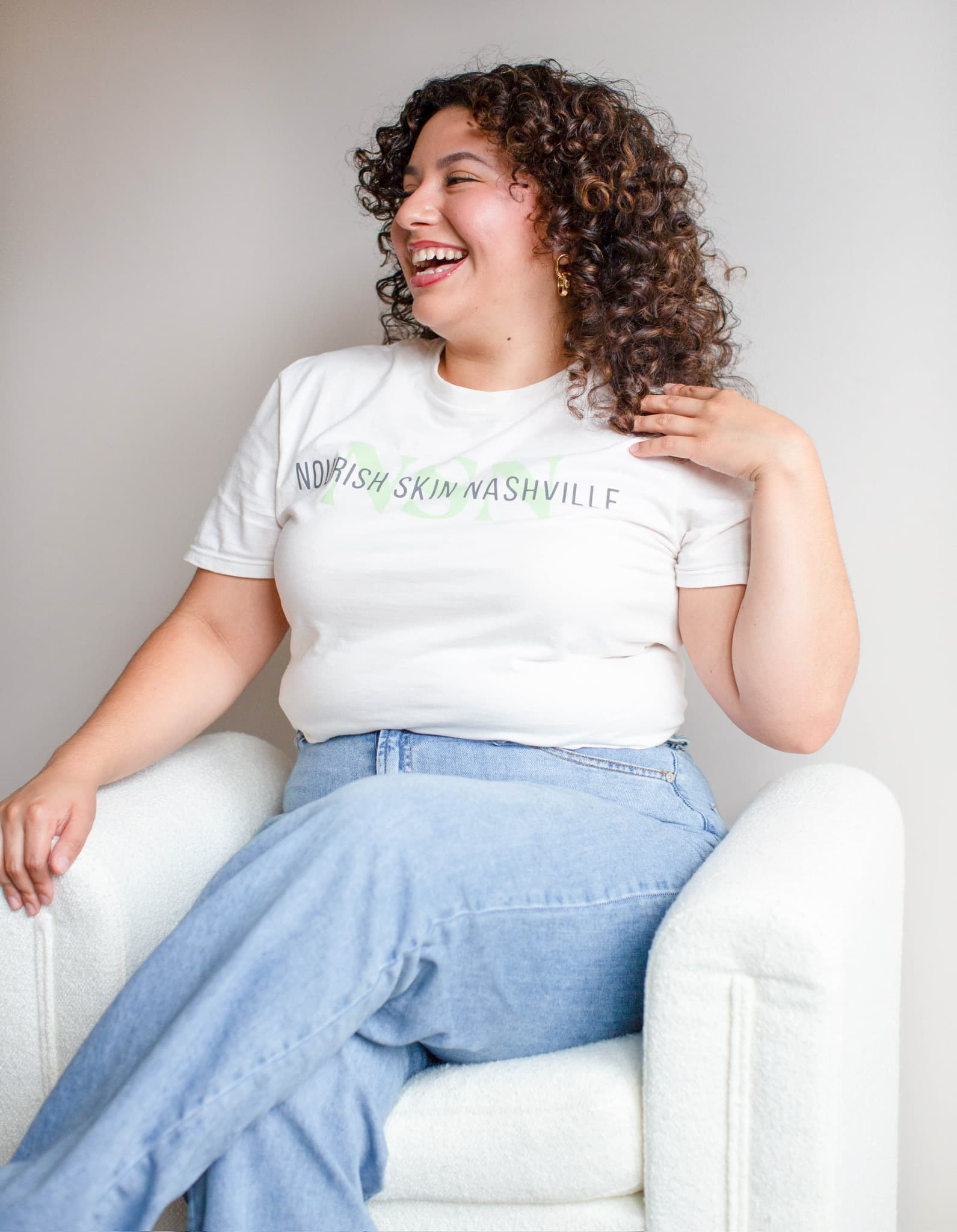 Roselyn Hernandez, licensed esthetician at Nourish Skin Nashville, seated in a white armchair, laughing with her eyes closed