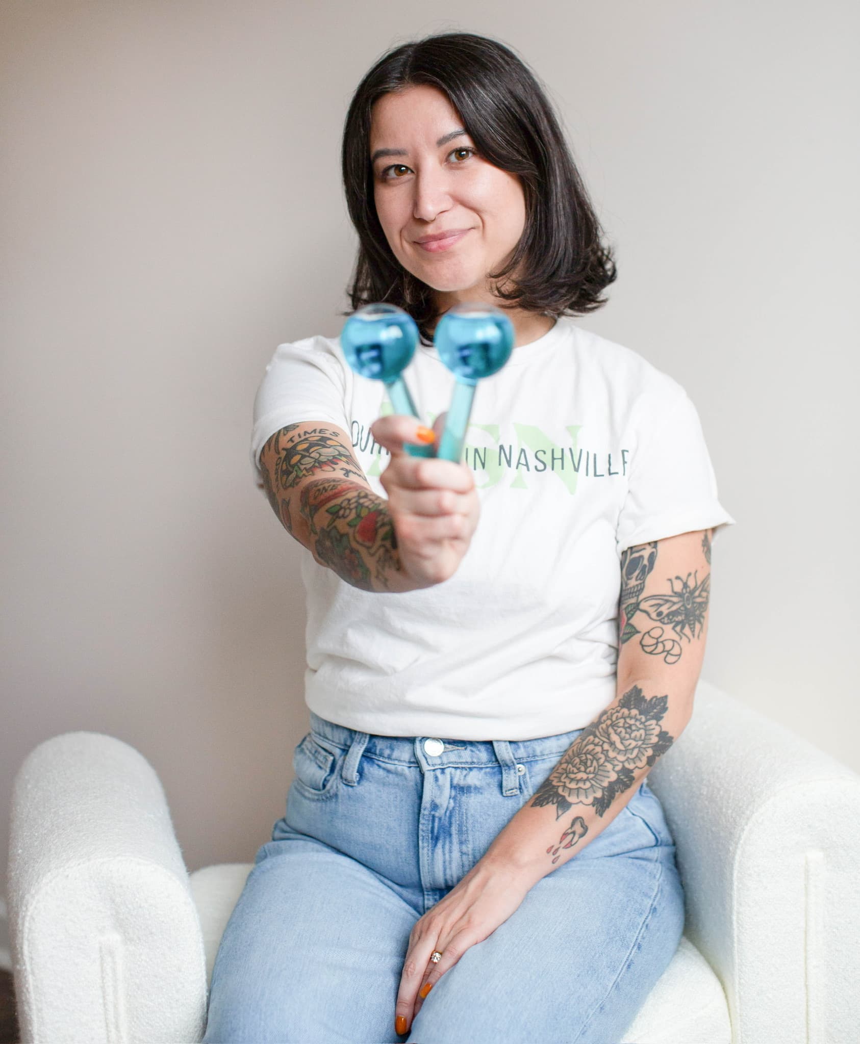 Sasha Park, licensed esthetician at Nourish Skin Nashville, seated in a white armchair, holding two blue ice globes towards the camera