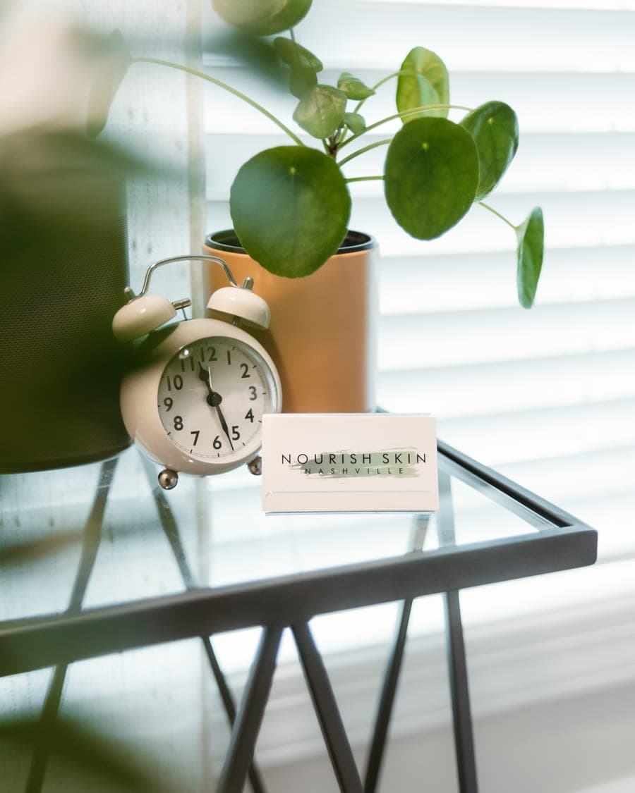 A Nourish Skin Nashville business card on a glass side table beside a plant and a clock, lit by a shuttered window