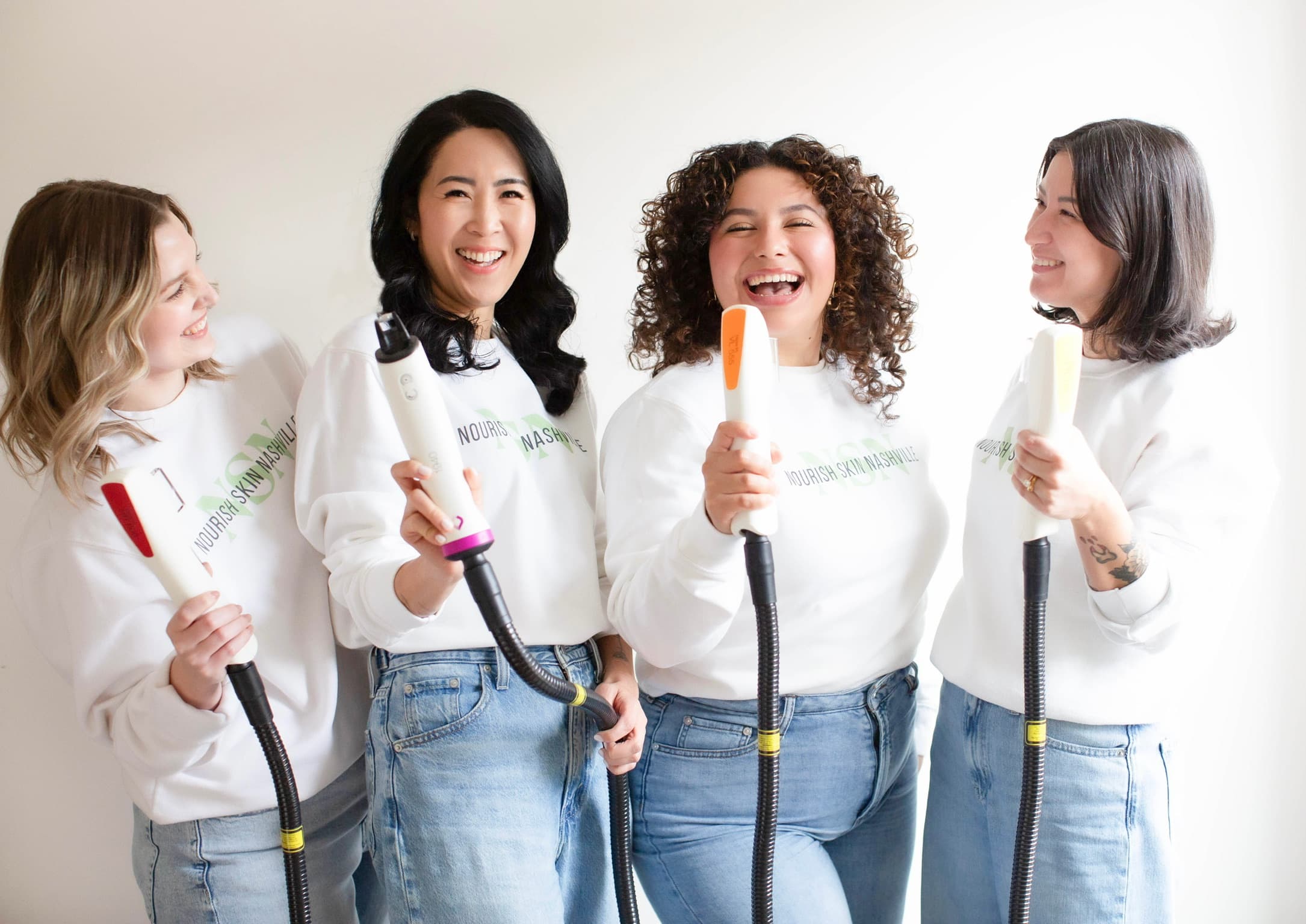 The four Nourish Skin Nashville estheticians laughing in a row, each holding a device handpiece up like a microphone