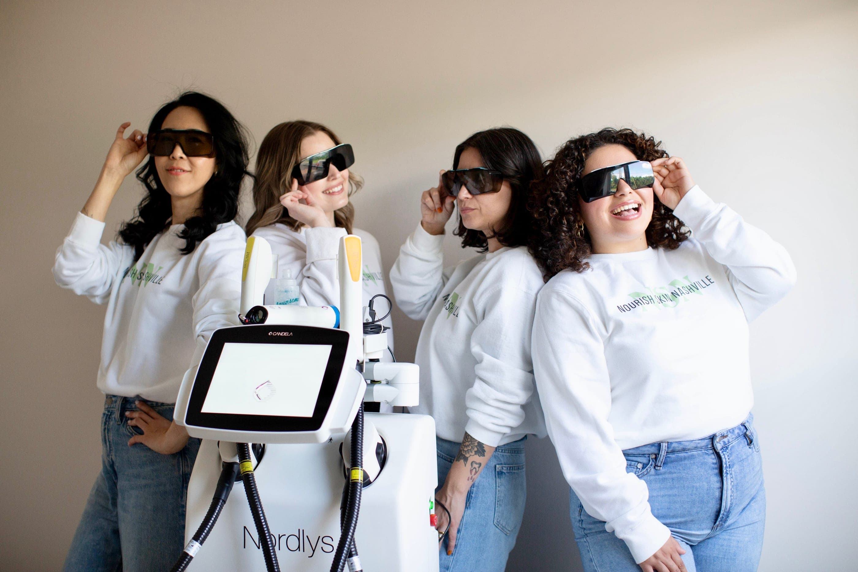 The four Nourish Skin Nashville estheticians in laser safety goggles around the studio's Candela Nordlys device, laughing